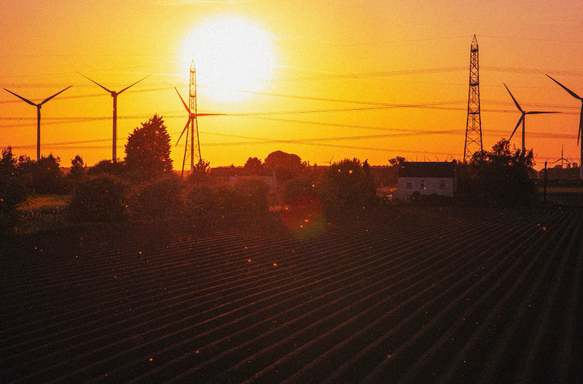 The sun setting behind wind turbines and pylons above a ploughed field with a farmhouse