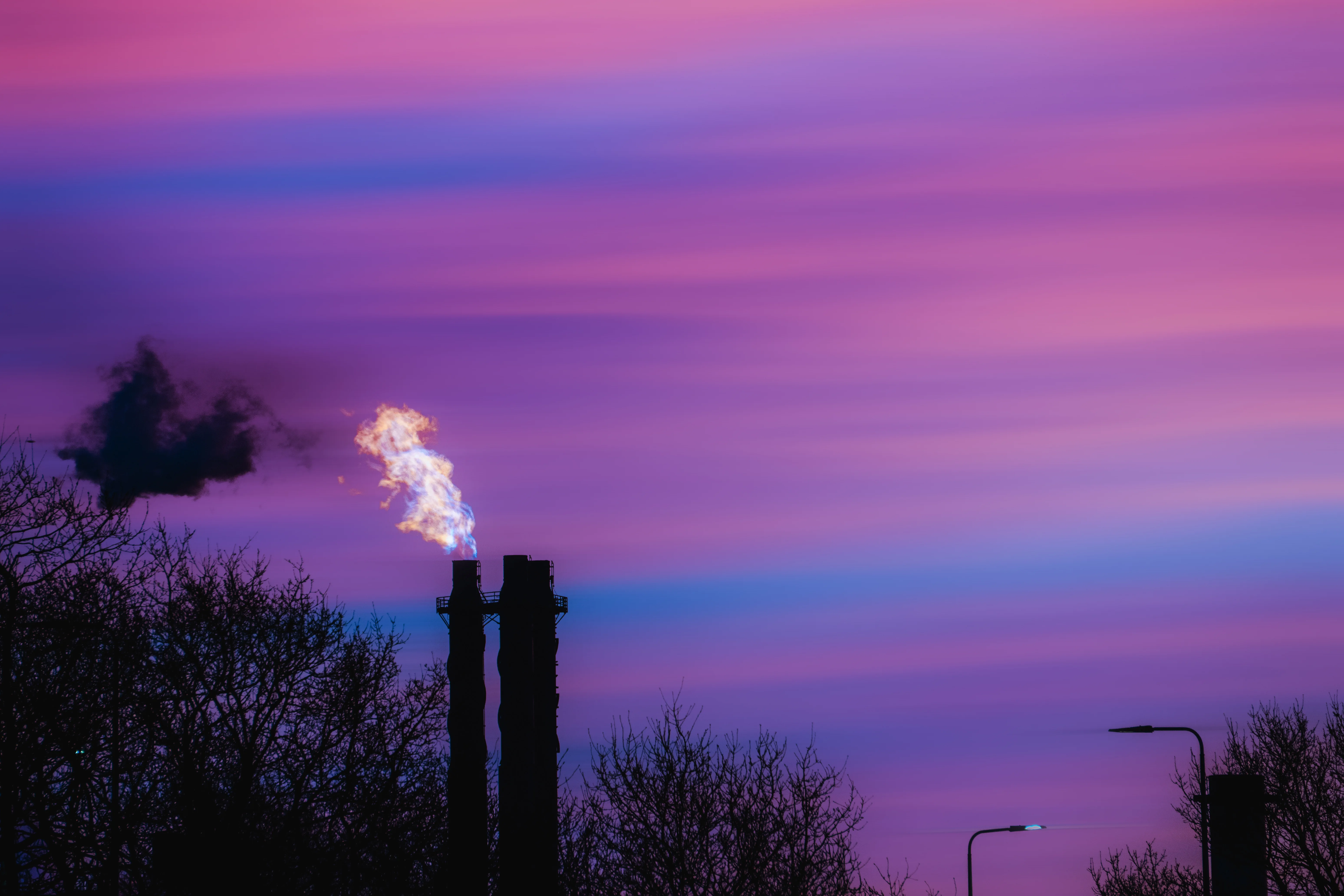 A flame rises from two industrial stacks against a violet evening sky