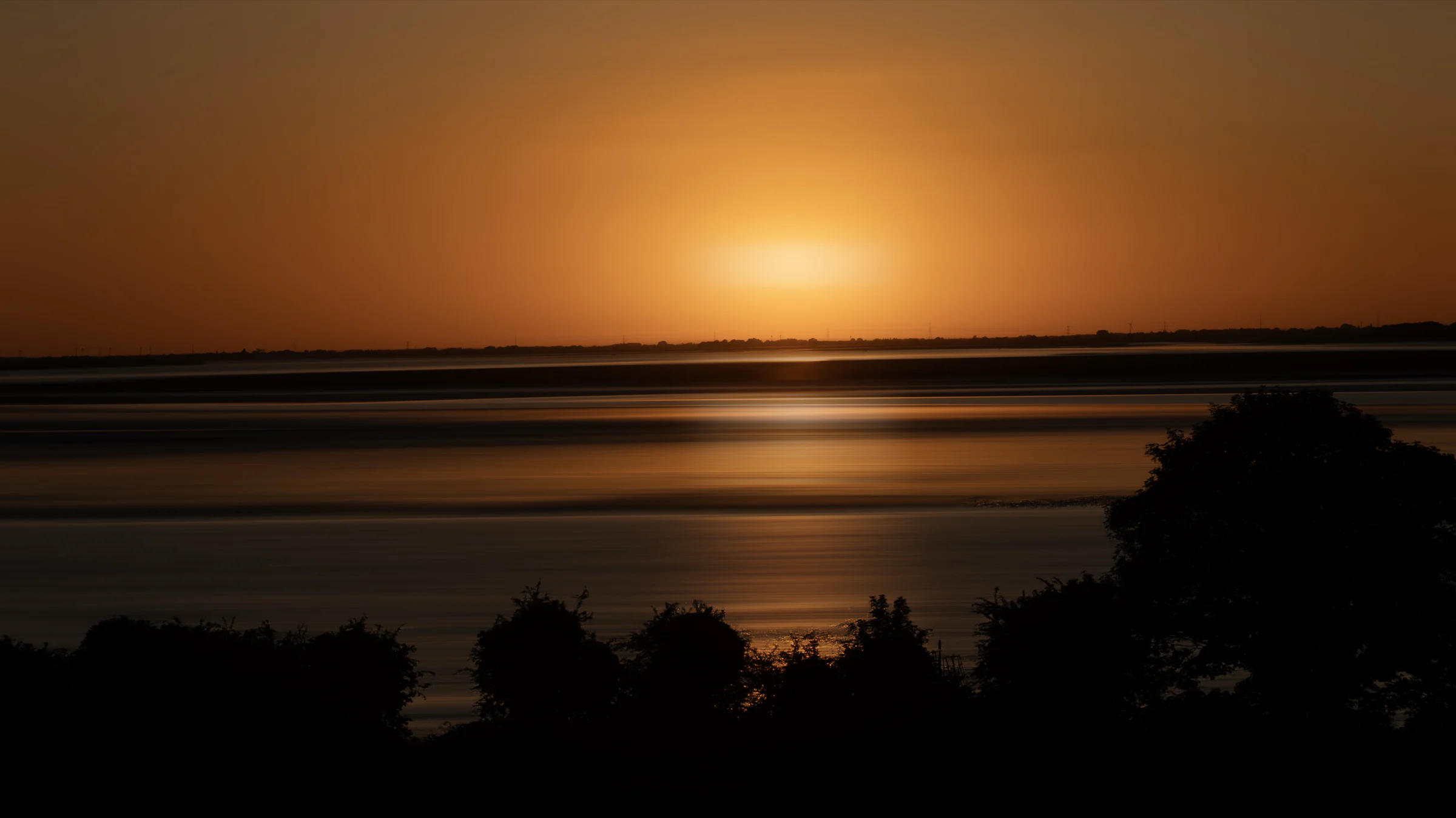 The sun low over water with silhouetted trees in the foreground